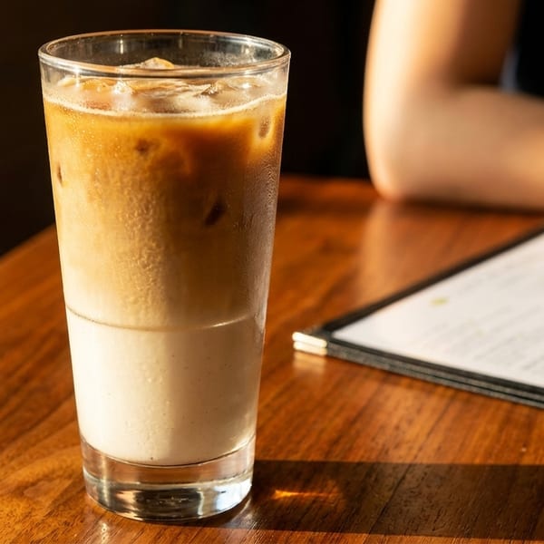 Iced latte on a wooden table with a menu and a person's arm in the background.
