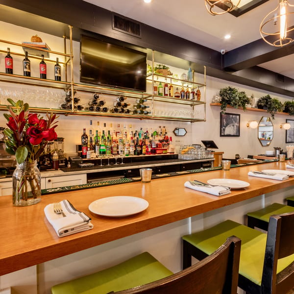 Well-stocked restaurant bar with wooden counter, green stools, liquor bottles, and gold shelving.