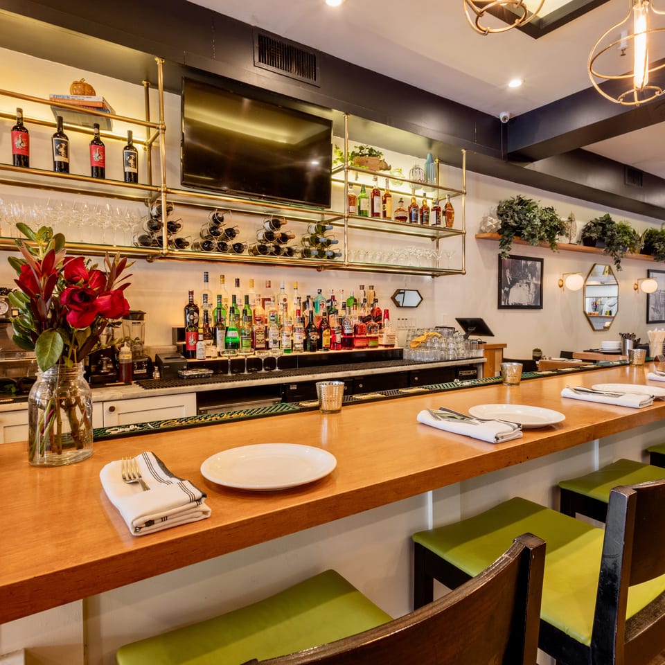Well-stocked restaurant bar with wooden counter, green stools, liquor bottles, and gold shelving.