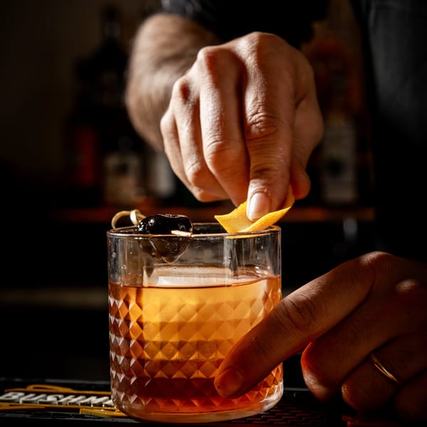 Bartender's hands garnishing a cocktail with an orange peel and a cherry on a dark bar.