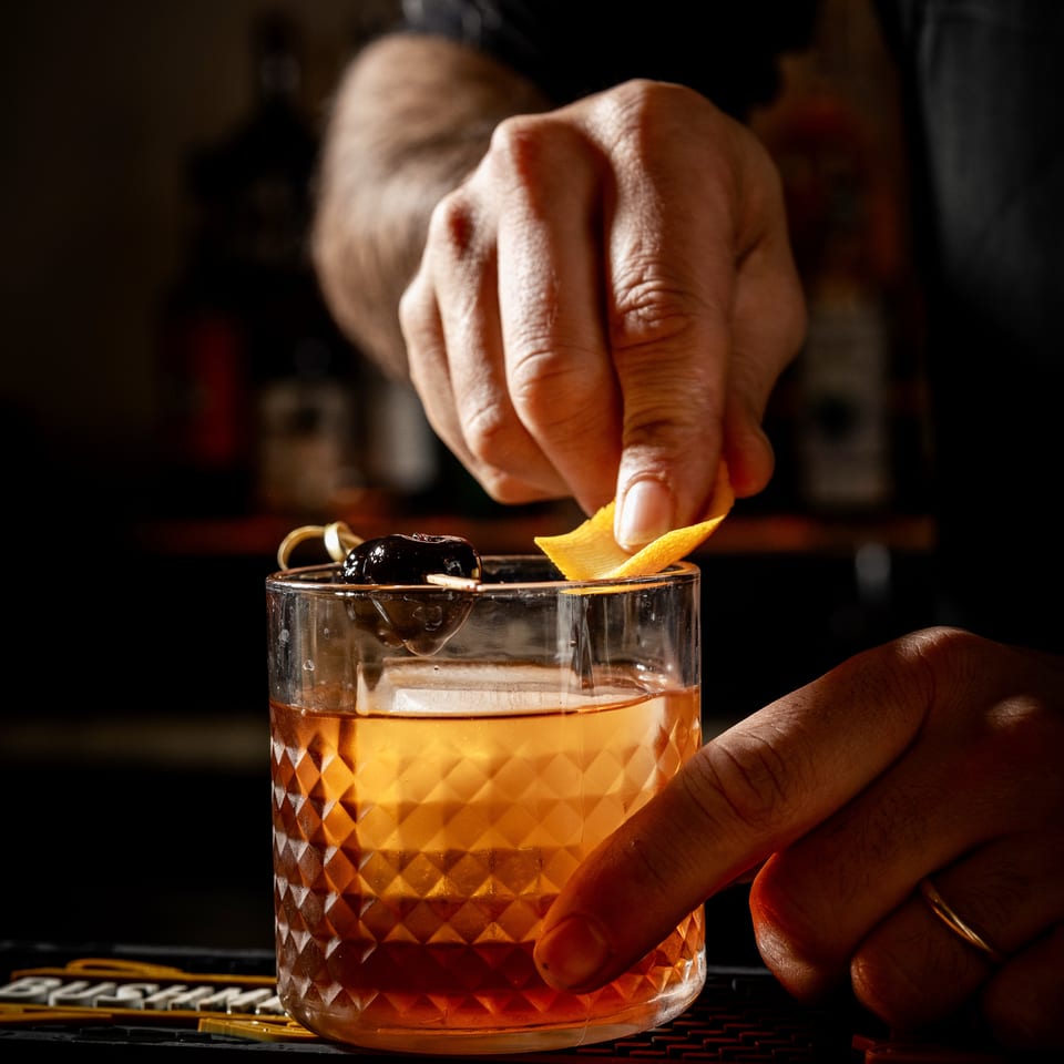 Bartender's hands garnishing a cocktail with an orange peel and a cherry on a dark bar.