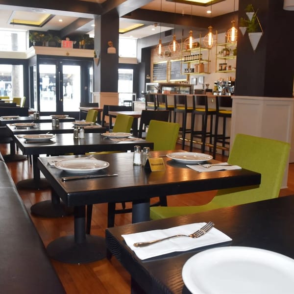 A modern restaurant interior with black tables, lime green chairs, and a bar area with gold shelving.