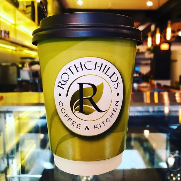 Rothchilds Coffee & Kitchen branded cup with a black lid, against a blurred cafe background.