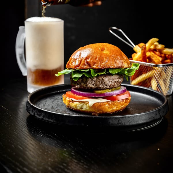 Hand pouring beer into a frosty mug next to a burger and a basket of golden fries.