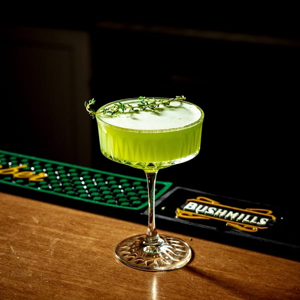 A vibrant green cocktail in a coupe glass, garnished with herbs, sits on a wooden bar.