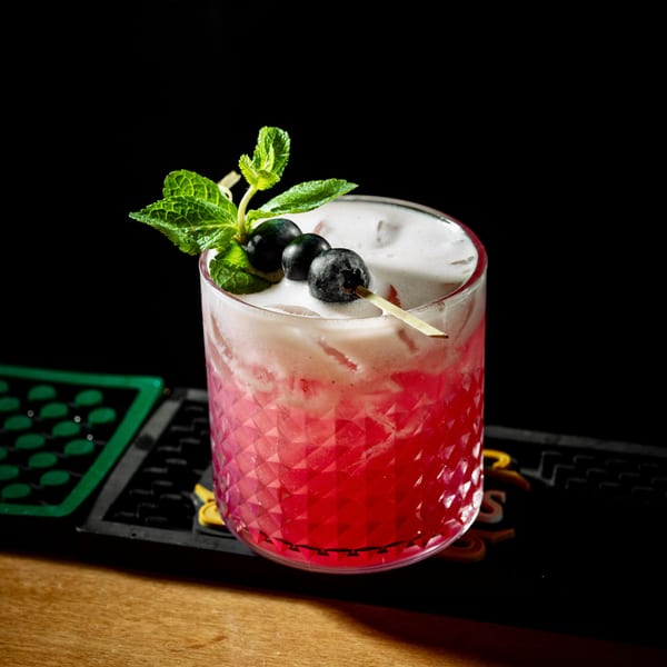Pink cocktail with white foam, garnished with blueberries and mint, on a wooden bar.