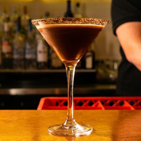 A chocolate martini with a sugared rim sits on a bar, with a bartender in the background.