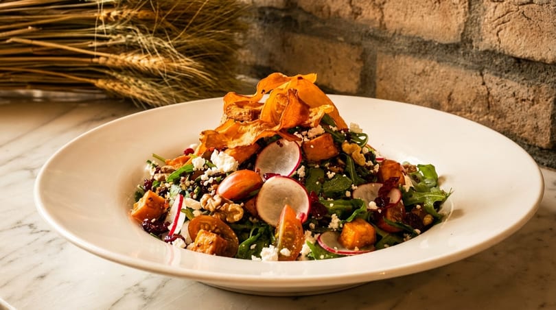 A vibrant salad with greens, radishes, tomatoes, feta, and sweet potato chips in a white bowl.