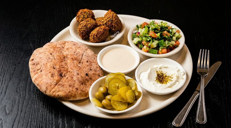 A platter of falafel, pita bread, salad, pickles, olives, tahini, and labneh on a dark table.
