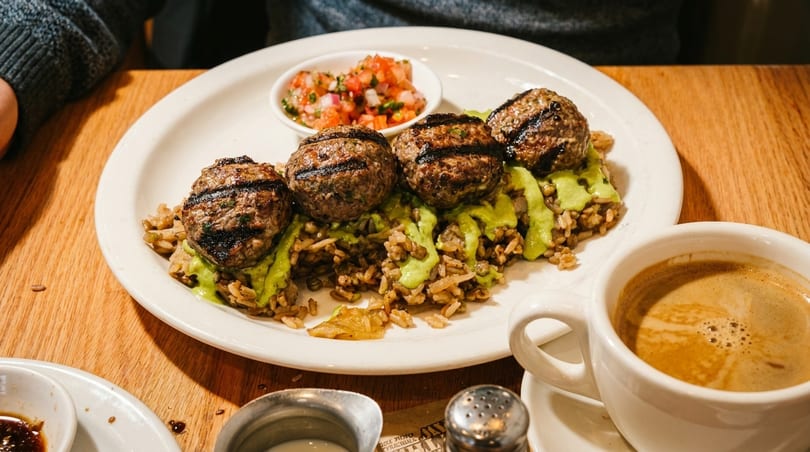 Grilled meatballs with green sauce over rice, salsa, and a cup of coffee on a wooden table.