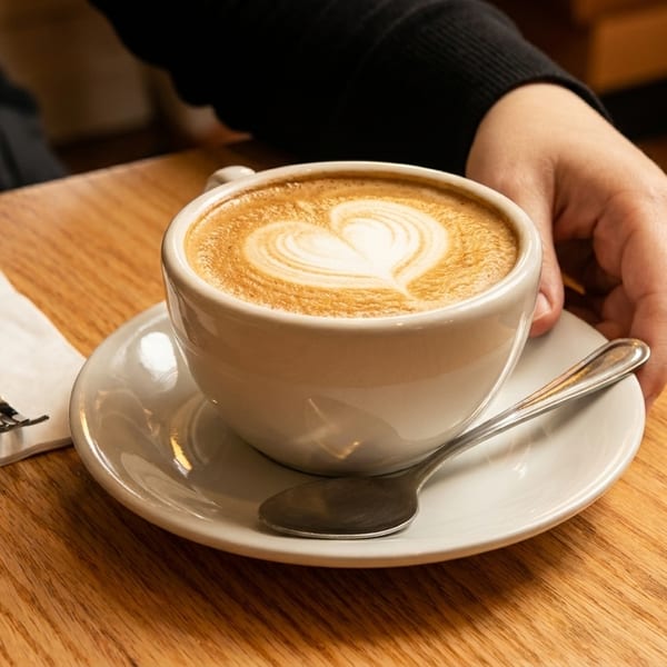 A person's hand rests next to a white cup of latte with heart-shaped foam art on a wooden table.